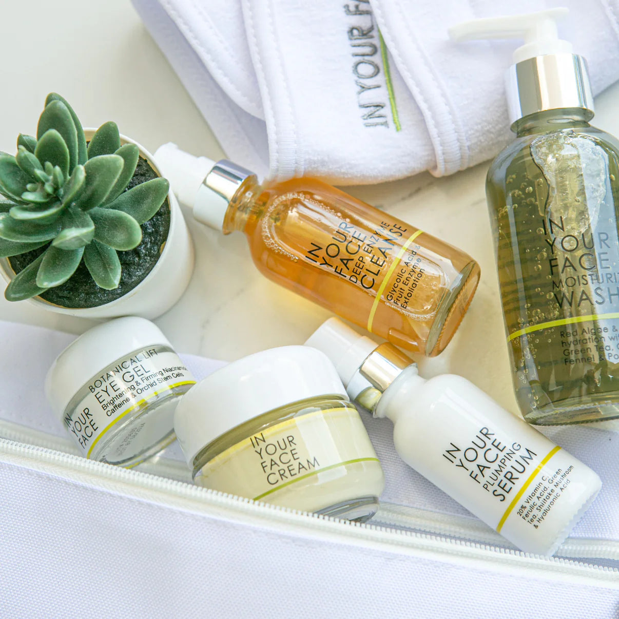 A collection of skincare travel kit products including moisturizers, exfoliators, serums, and cleansing products, displayed alongside a small potted plant and a towel with text on it.