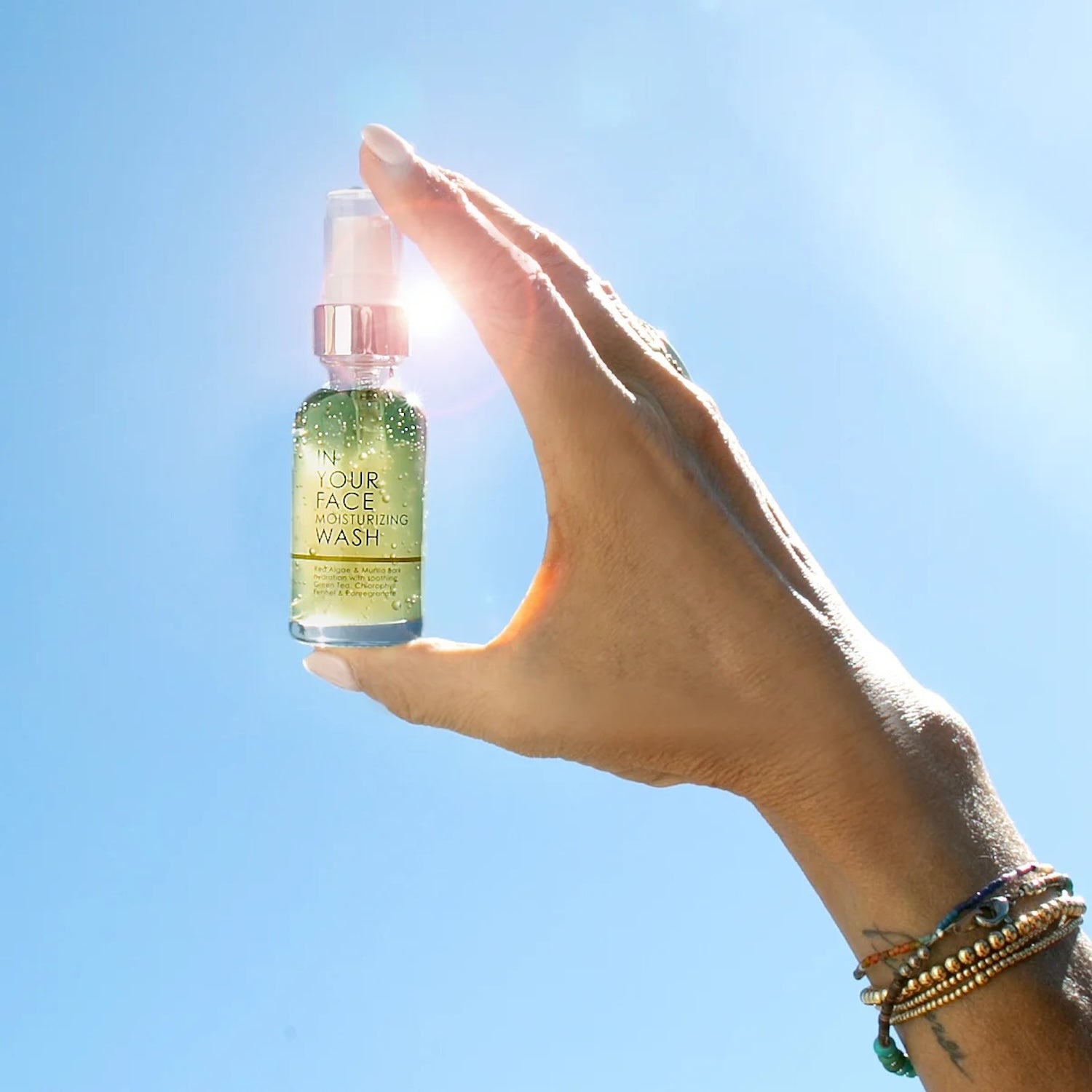 In Your Face Moisturizing Wash Mini held against a bright blue sky, highlighting its travel size and lightweight, refreshing formula for glowing skin anywhere.