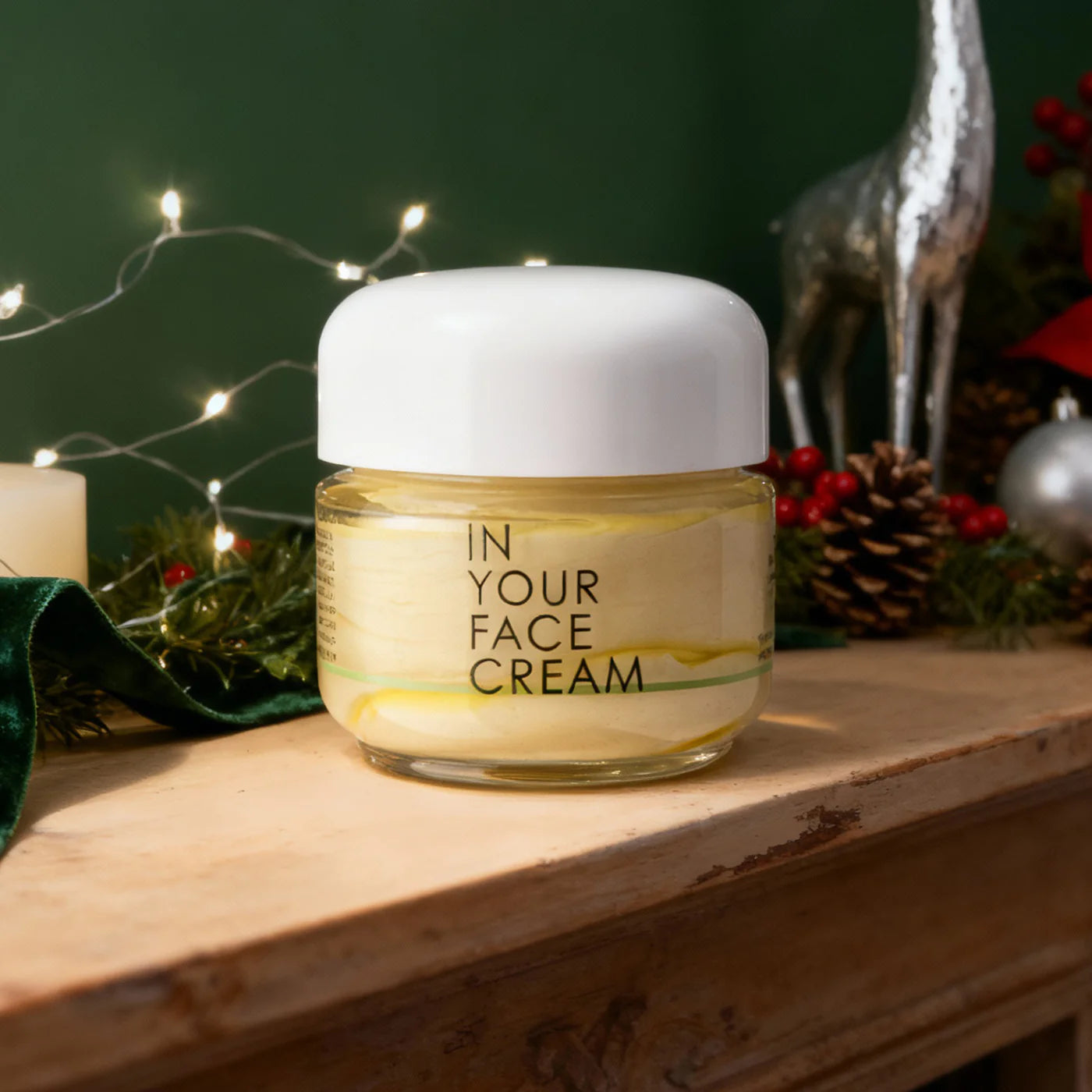 Jar of 'In Your Face Cream' on a wooden surface with festive decorations in the background