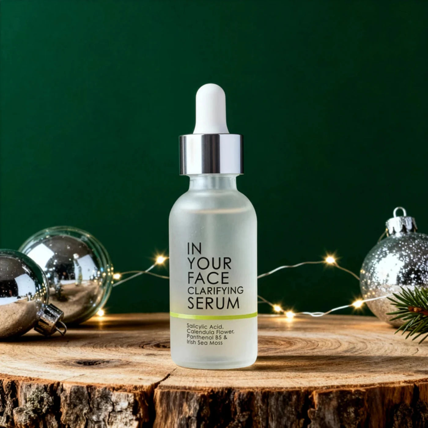 Bottle of 'In Your Face Clarifying Serum' on a wooden surface with Christmas ornaments in the background.