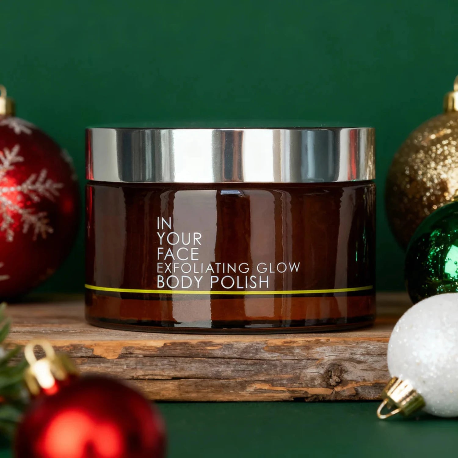 Jar of 'In Your Face Exfoliating Glow Body Polish' with Christmas ornaments on a green background