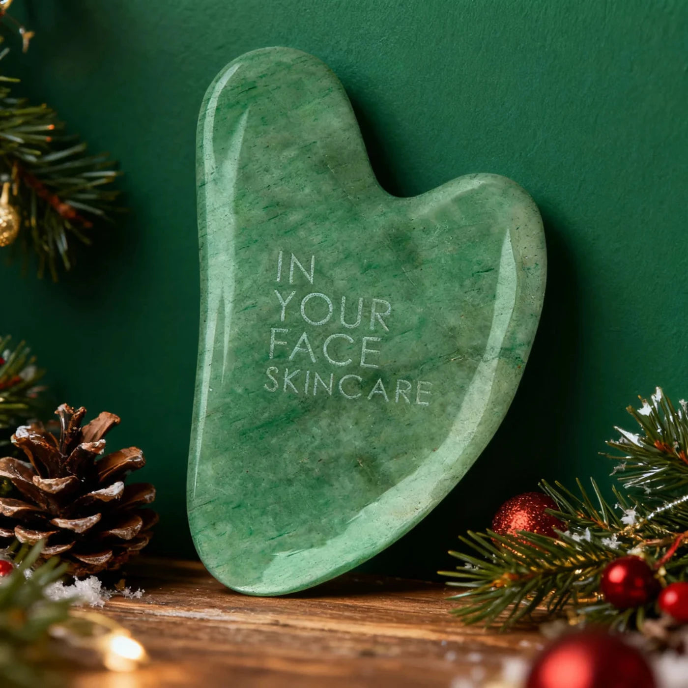 Green gua sha tool with 'IN YOUR FACE SKINCARE' text on a festive background with pine cones and decorations.