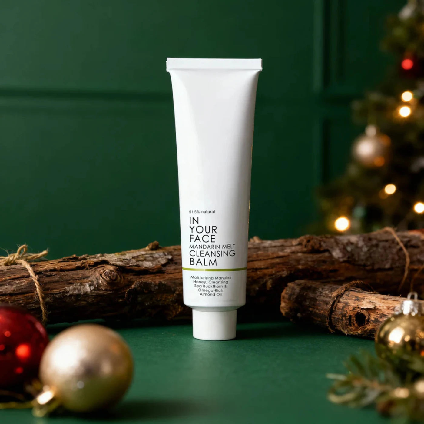 Tube of 'In Your Face' cleansing balm on a green background with Christmas decorations.