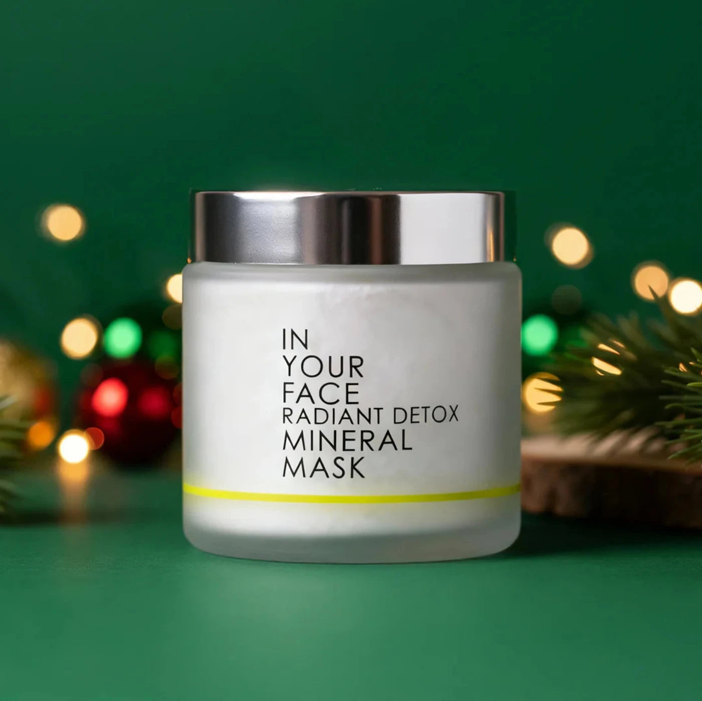 Jar of 'In Your Face Radiant Detox Mineral Mask' on a green background with festive lights.