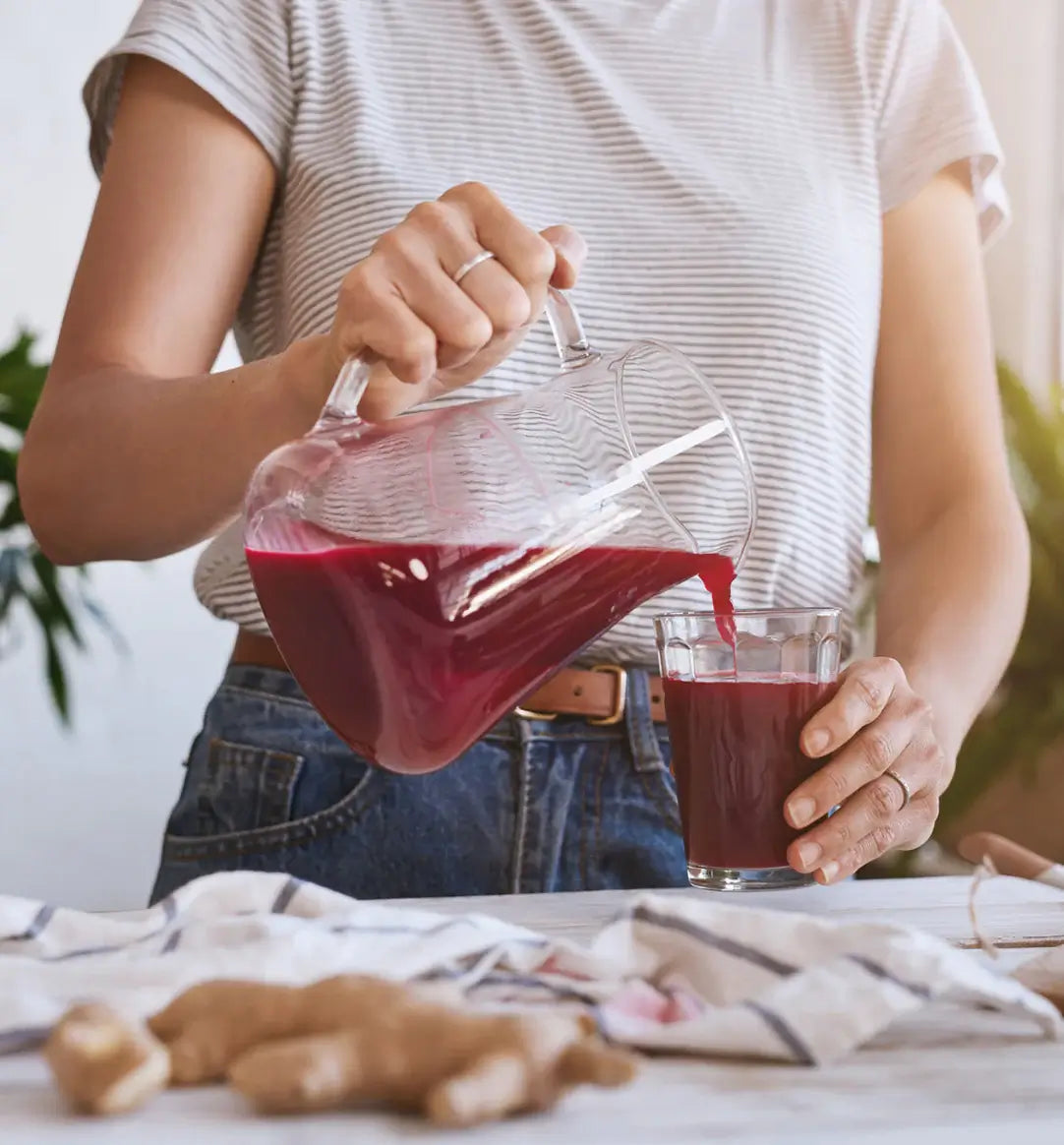 Woman pouring fresh red juice from a glass pitcher into a cup on a kitchen table with ginger root in the foreground