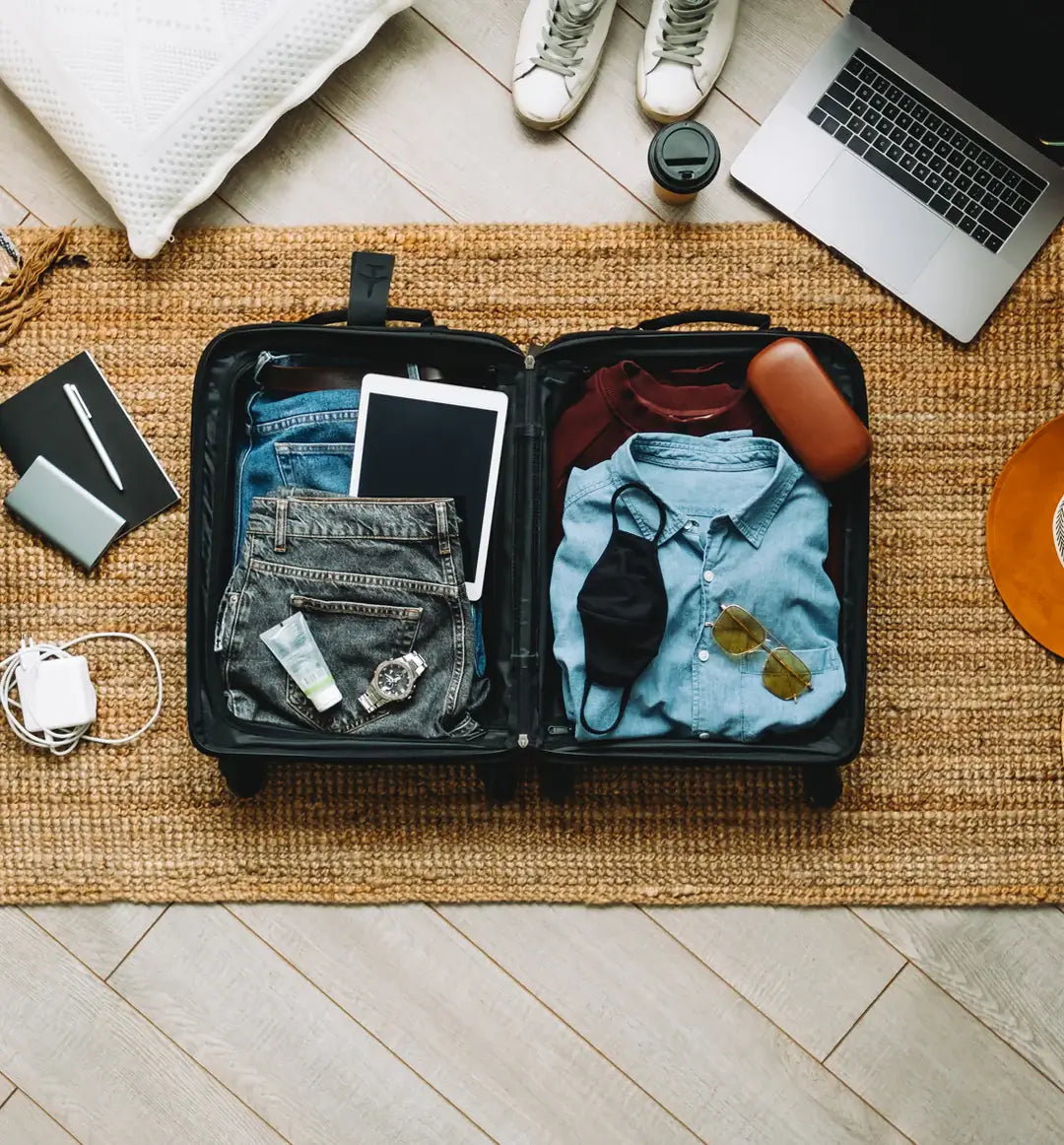 Open suitcase packed with clothes, tablet, sunglasses, watch, and travel essentials on a woven rug next to laptop and coffee cup – ready for a trip