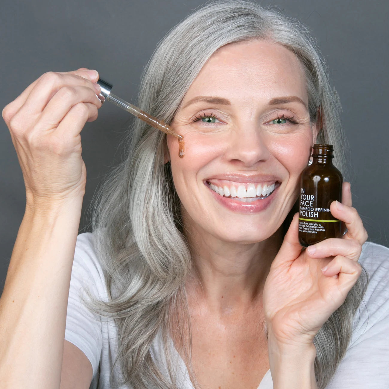 Woman applying skincare product with dropper and bottle against a gray background
