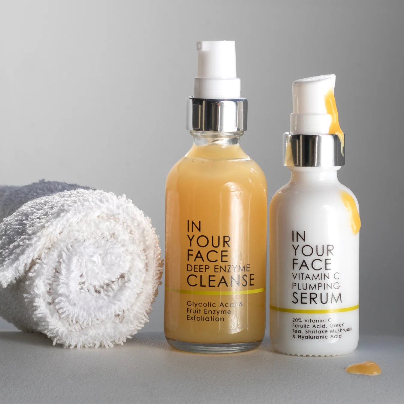 Two skincare bottles labeled 'Deep Enzyme Cleanse' and 'Vitamin C Plumping Serum' with a towel on a gray background.