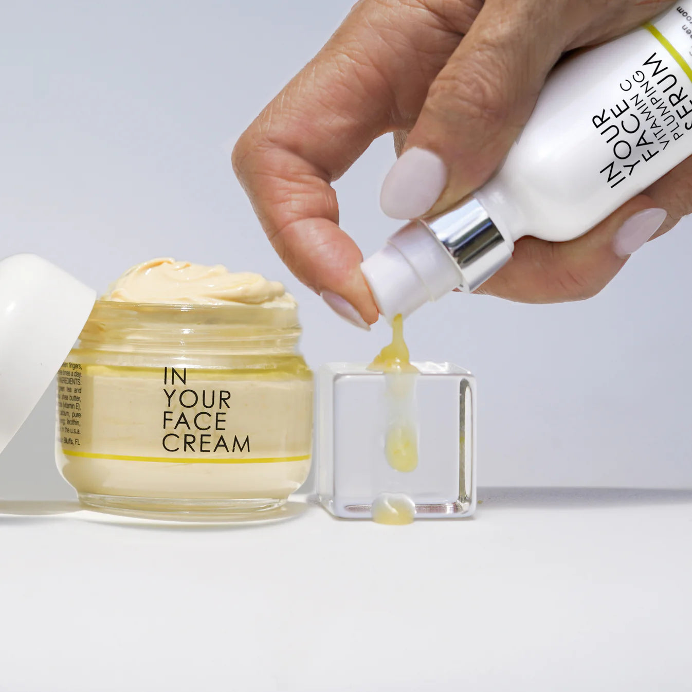 Hand pouring cream from a bottle into a small container next to 'In Your Face Cream' jar on a light background