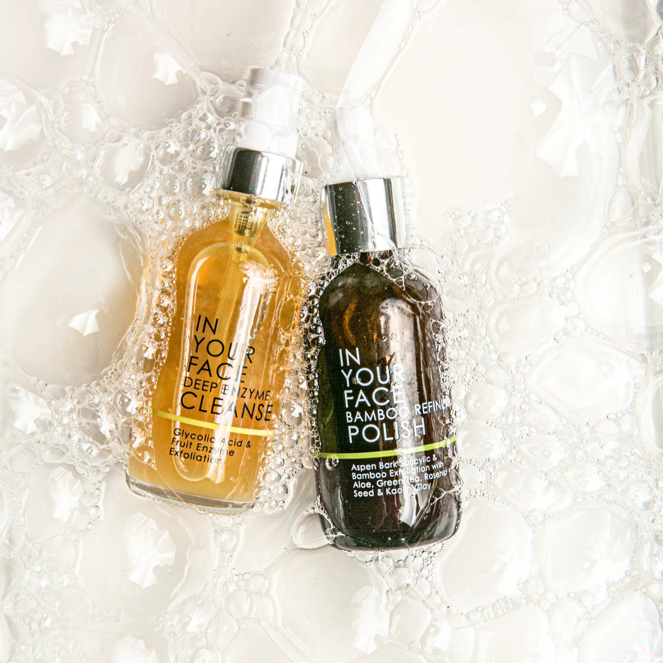 Two bottles of skincare products, 'In Your Face Deep Enzyme Cleanse' and 'Bamboo Refining Polish', surrounded by bubbles.