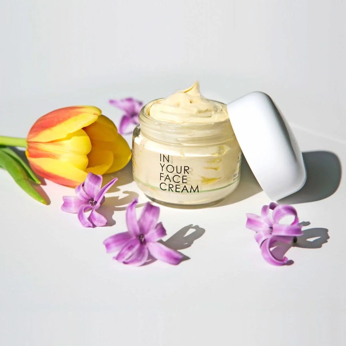 In Your Face Cream jar with open lid surrounded by a yellow tulip and purple flowers – hydrating natural face moisturizer