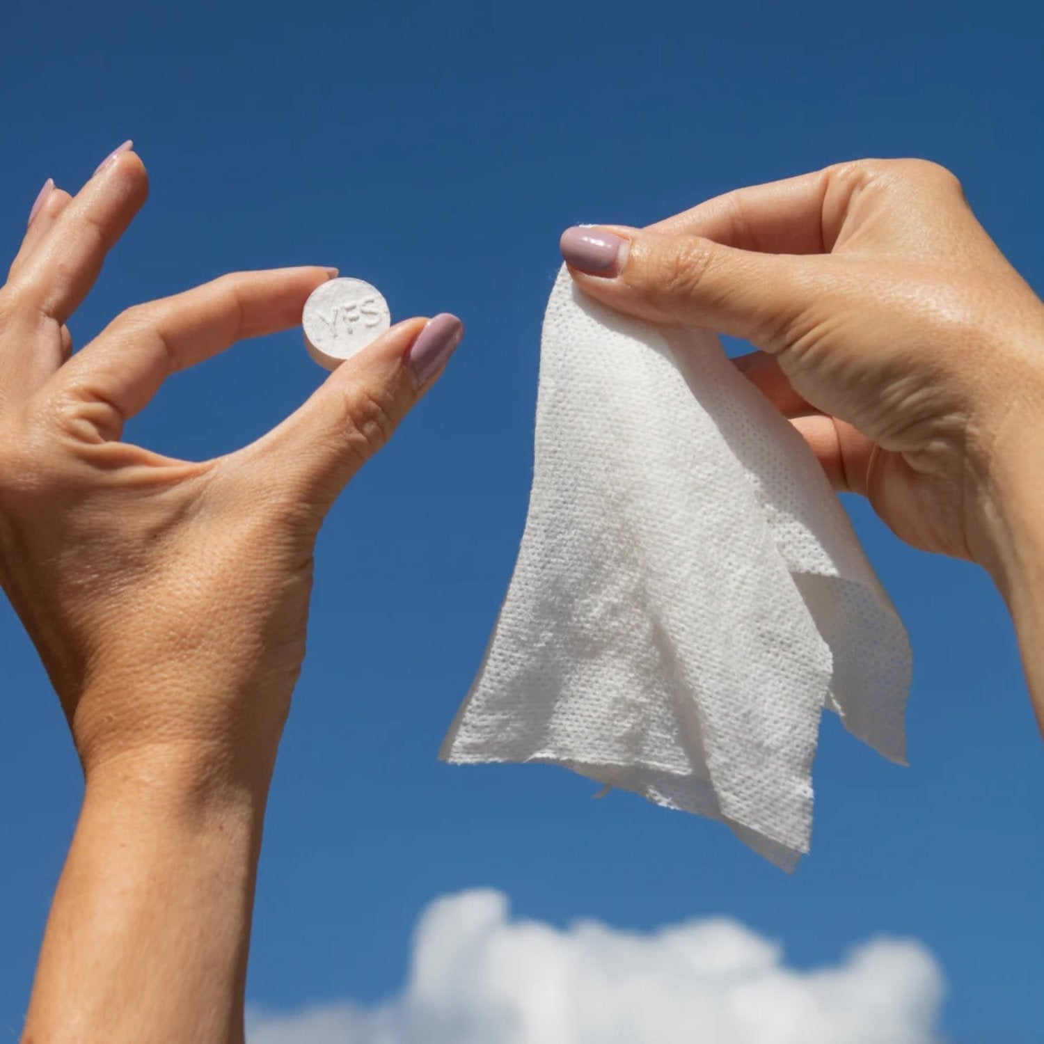 Hands holding a compressed In Your Face Cleansing Cloth and its expanded form against a blue sky, demonstrating the product’s biodegradable and reusable design.