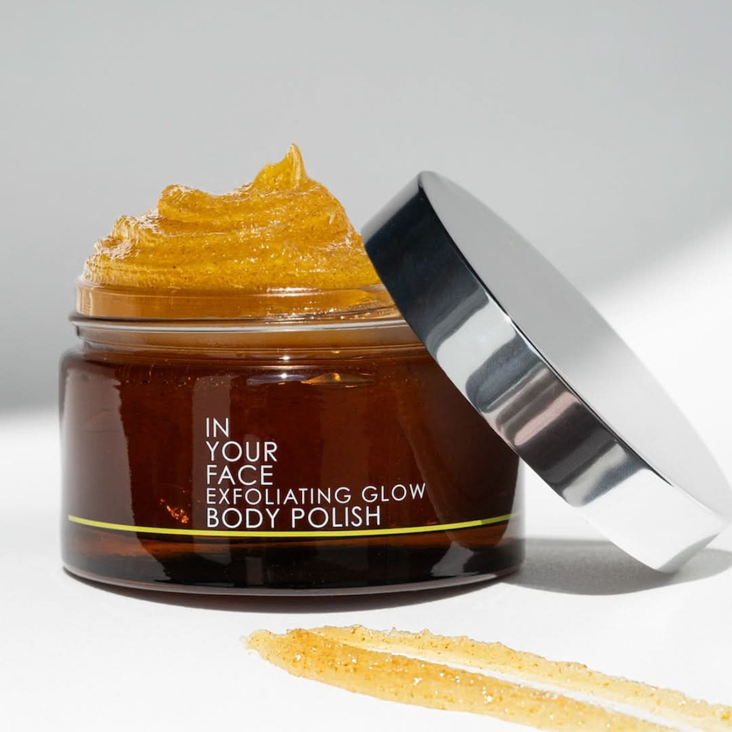 Open jar of In Your Face Exfoliating Glow Body Polish with its thick golden texture and a swatch on a clean white surface, showcasing its exfoliating formula.