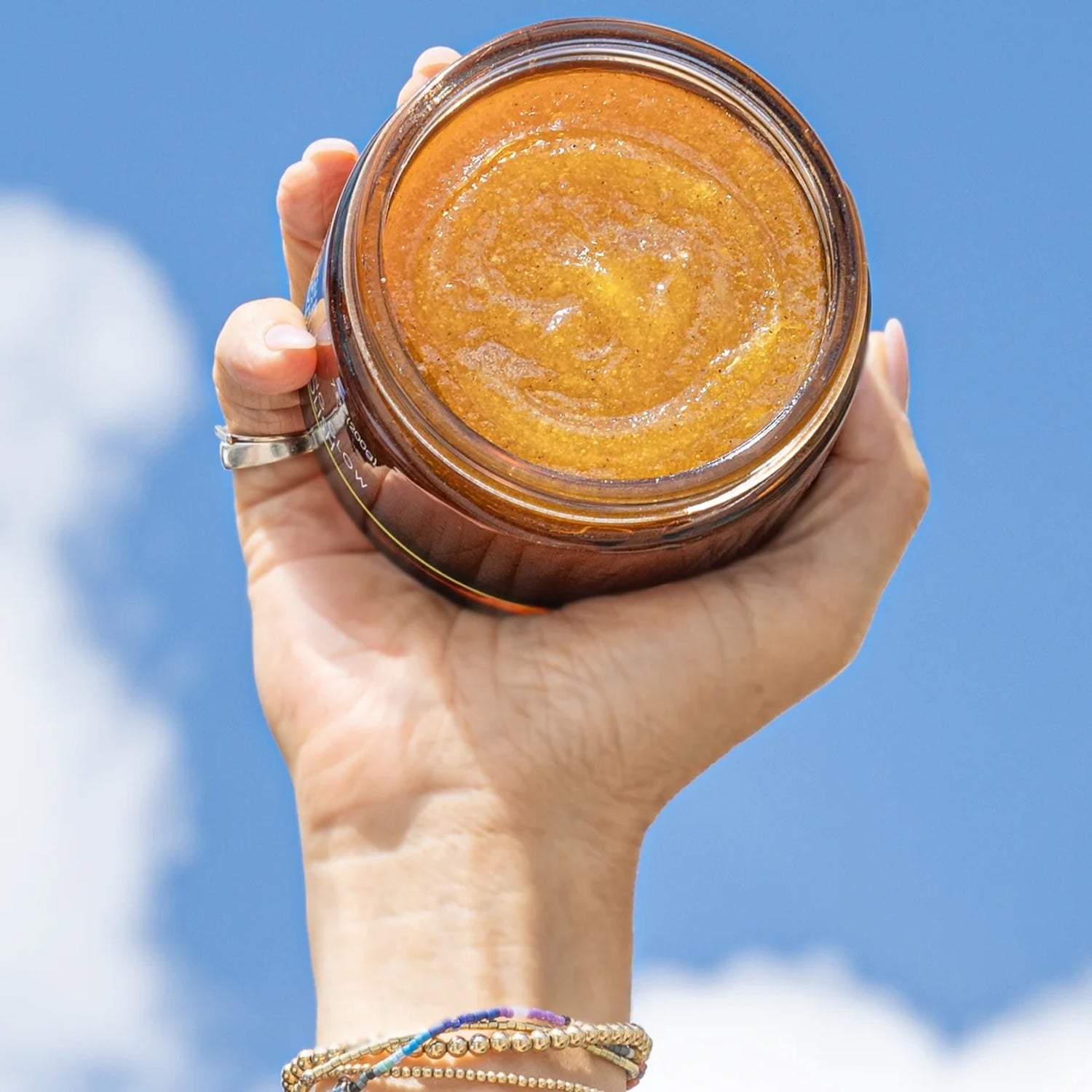 Jar of In Your Face Exfoliating Glow Body Polish held up against a bright blue sky, emphasizing its skin-brightening and hydrating benefits.