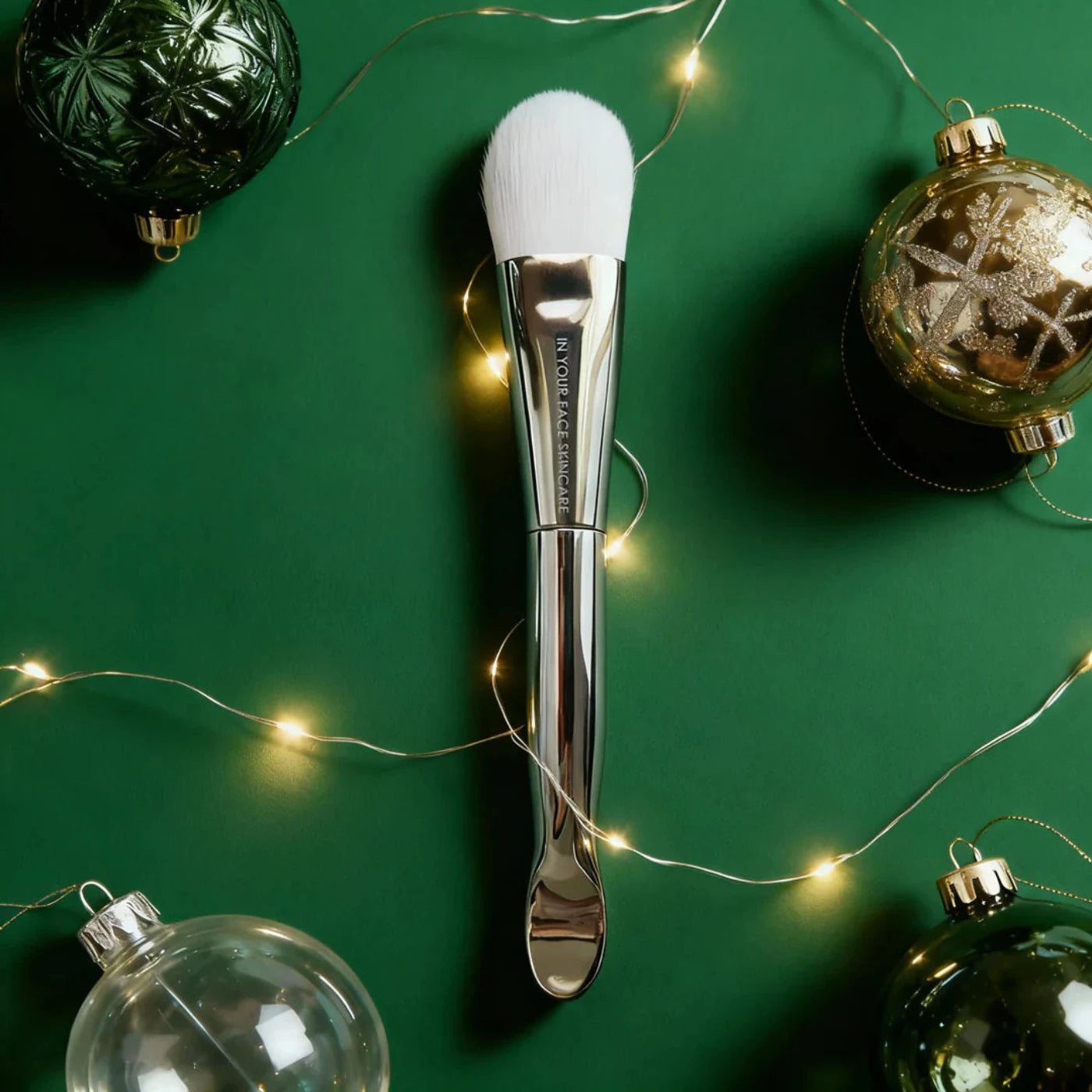 Makeup brush with silver handle on a green background with Christmas ornaments and string lights