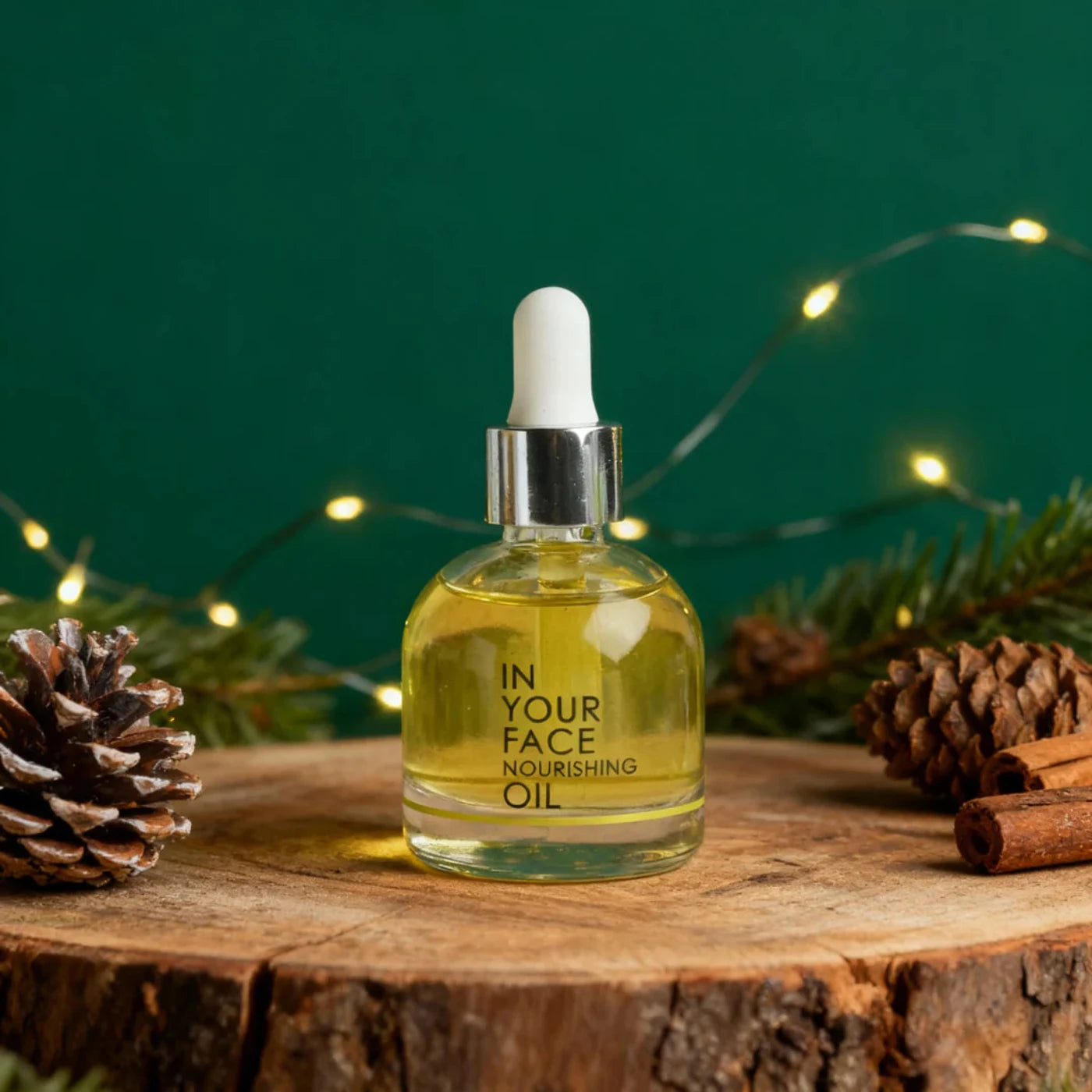 Bottle of 'In Your Face Nourishing Oil' on a wooden surface with pine cones and lights in the background.