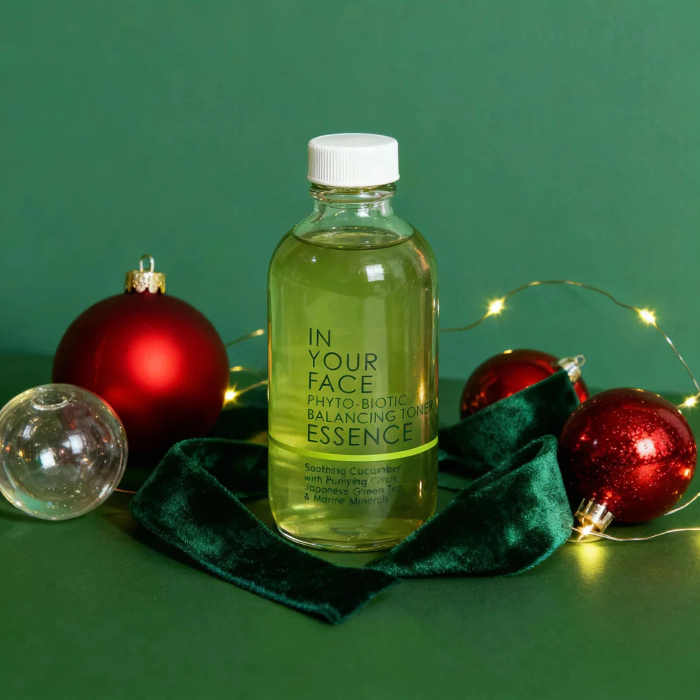 Bottle of 'In Your Face' toning essence with Christmas ornaments on a green background