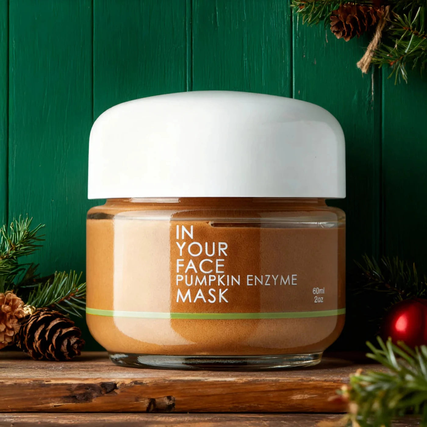 Jar of 'In Your Face Pumpkin Enzyme Mask' with festive decorations on a wooden surface and green background.