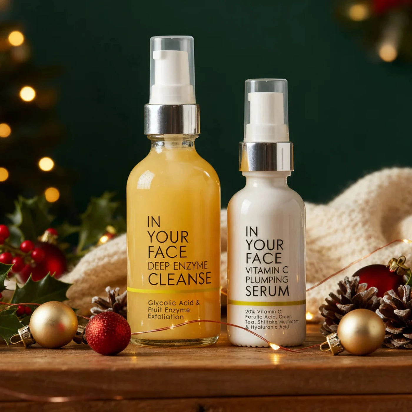 Two skincare bottles, 'In Your Face Deep Enzyme Cleanse' and 'Plumping Serum', on a wooden surface with festive decorations.