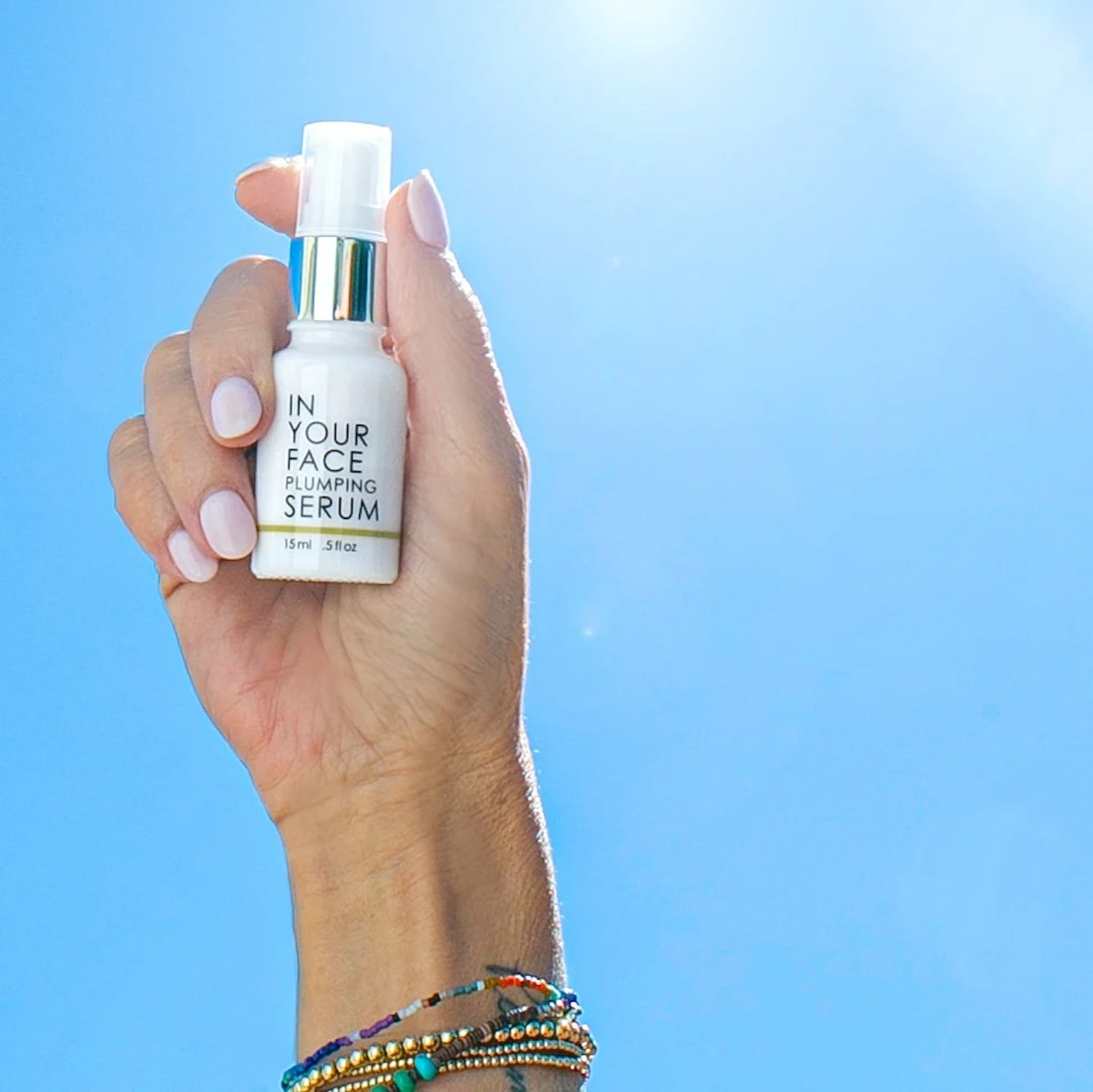 In Your Face Vitamin C Plumping Serum Mini held against a bright blue sky, showcasing its lightweight, travel-friendly design and skin-enhancing benefits.