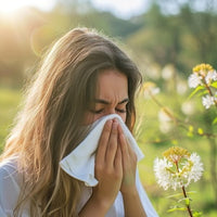 A woman outdoors in a sunny, blurred garden setting, holding a white tissue to her nose. She has long light brown hair and is wearing a white top.
