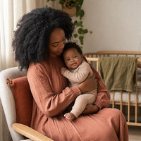 A woman with dark curly hair sits in a chair holding a baby in a softly lit nursery. The woman wears a dusty terracotta long-sleeve top and matching skirt, while the baby wears a light cream outfit.