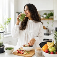 A woman in a white blouse stands in a bright kitchen, drinking a green smoothie from a glass. Bowls of fruit and vegetables, a blender, and fresh produce are arranged on the counter in front of her.