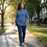 A woman walking on a sunlit tree-lined path wearing a slate blue long-sleeve top and dark navy pants.