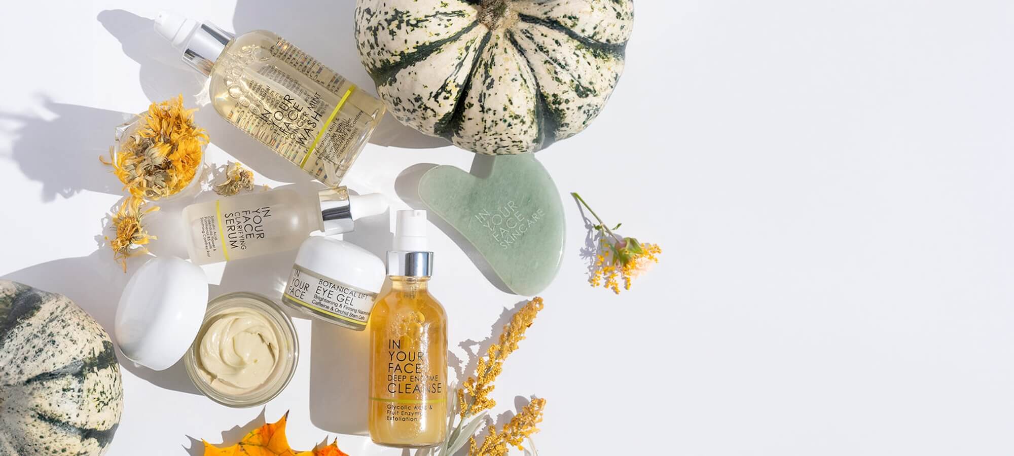 Autumn skincare essentials arranged on a white background, featuring botanical serums, creams, and a jade gua sha tool, surrounded by dried flowers and a decorative gourd.