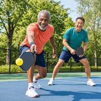 Two men playing pickleball on an outdoor court, one in an orange shirt and one in a teal shirt, holding paddles and smiling.