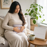 A woman in a cozy living room setting, sitting on a couch wrapped in a cream knit sweater and blanket, holding a light purple belly support pillow. A small wooden table next to her holds a mug and a monthly calendar, with a green potted plant in the background.